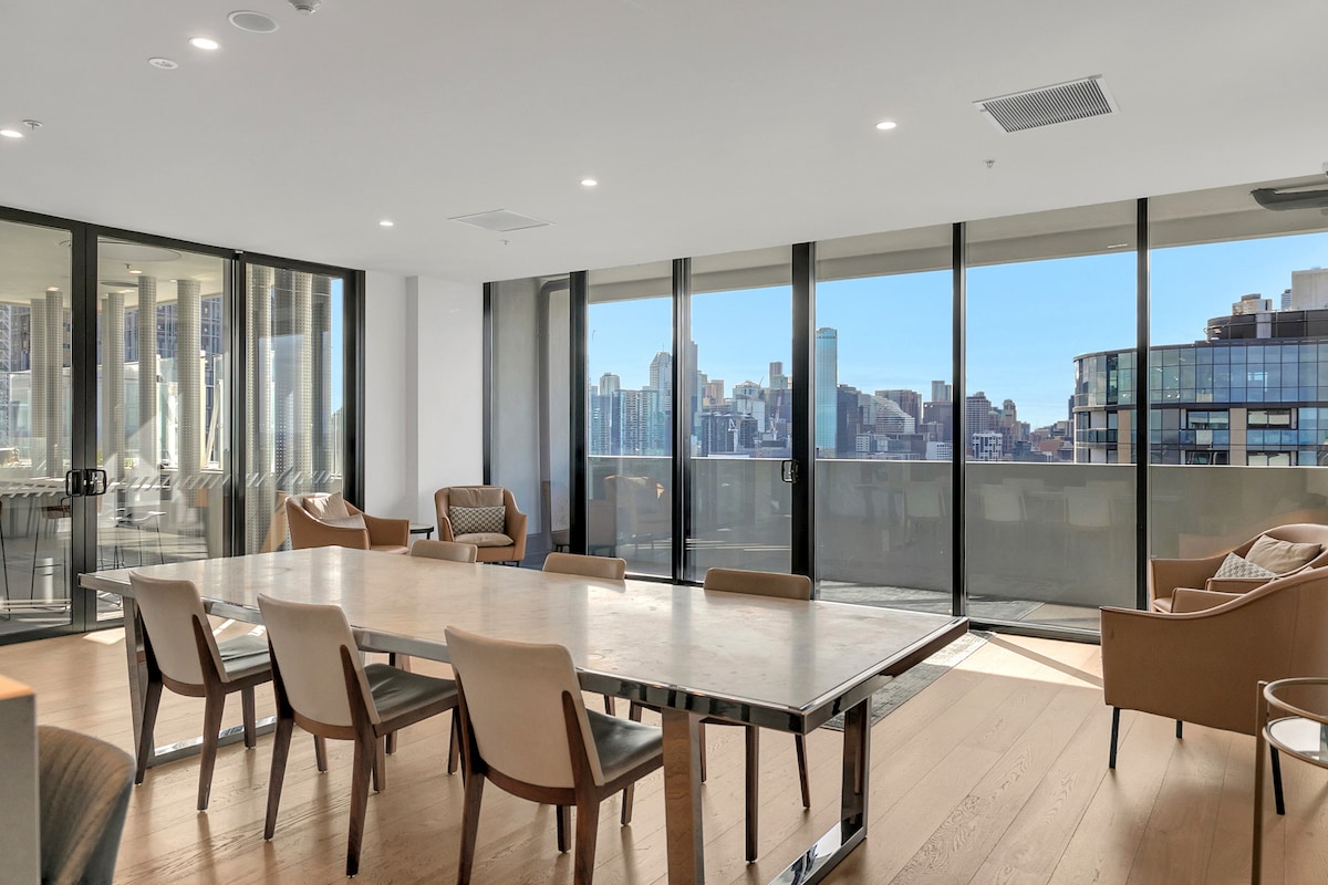 The open-plan living area is bathed in natural light, featuring a large dining table surrounded by beige chairs. Floor-to-ceiling glass doors provide access to a private balcony with views of the city skyline, enhancing the spacious feel of the room.