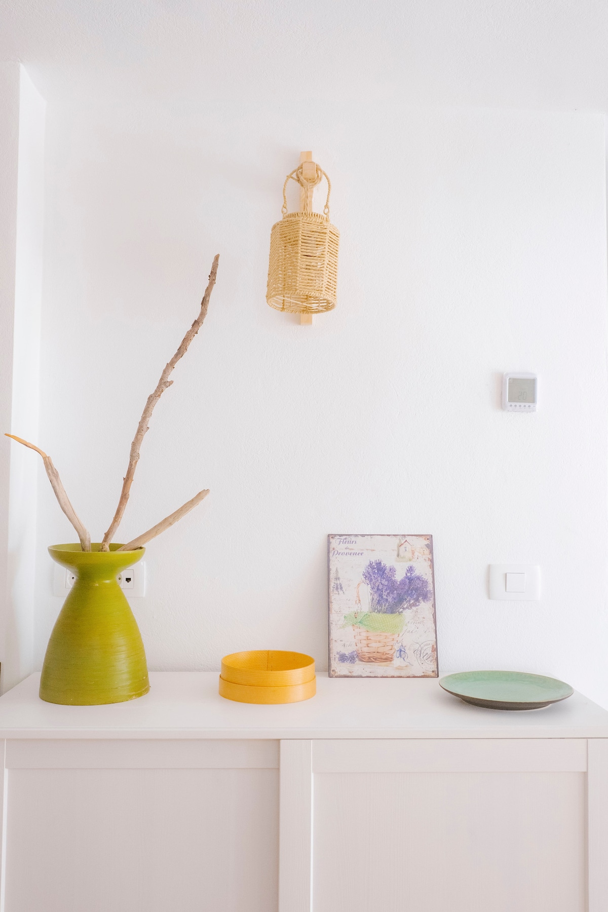 A shelf displays a green vase with branches, a yellow bowl, and a decorative plate. A framed art piece featuring a floral arrangement is positioned nearby. A woven lantern is mounted on the wall above, contributing to a cozy, inviting environment.