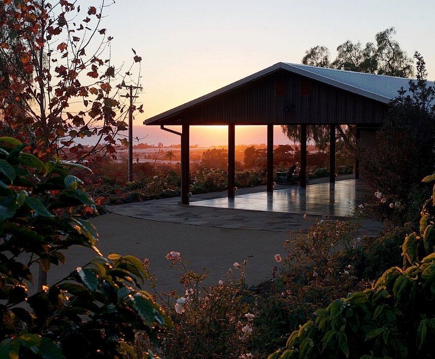 The image captures a modern structure with a spacious canopy, situated among lush greenery. The setting sun casts a warm glow, illuminating the surrounding garden filled with flowering plants. The tranquil landscape suggests a peaceful outdoor space for relaxation or gathering.