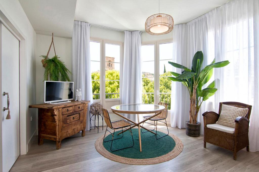A bright living area features large windows that provide views of greenery outside. A round dining table surrounded by four chairs sits on a textured rug. A potted plant and a vintage sideboard enhance the space, complemented by natural light filtering through sheer curtains.