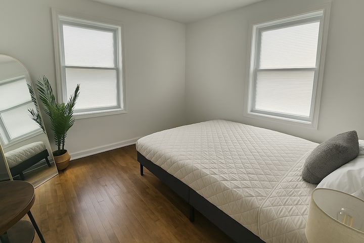 Quarto minimalista e iluminado com uma cama queen aconchegante, roupa de cama acolchoada macia, espelho de corpo inteiro, vaso de plantas e pisos de madeira natural. Um espaço tranquilo e limpo, perfeito para noites tranquilas ou estadias mais longas.