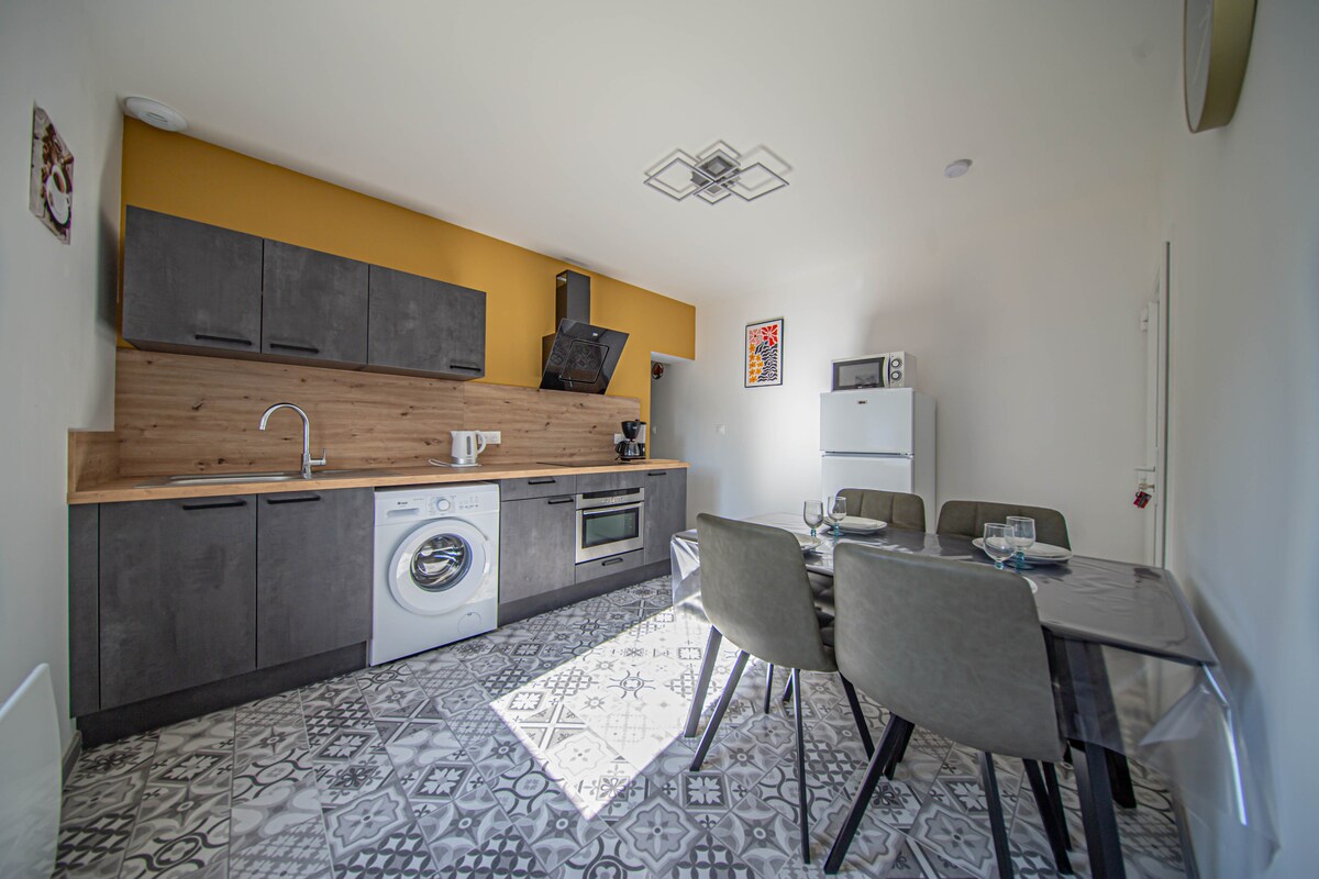 A modern kitchen is displayed, featuring sleek cabinetry in dark tones and a warm wooden accent wall. Essential appliances include a washing machine, microwave, and refrigerator. A dining table with grey chairs is set for meals, illuminated by natural light filtering through the space.