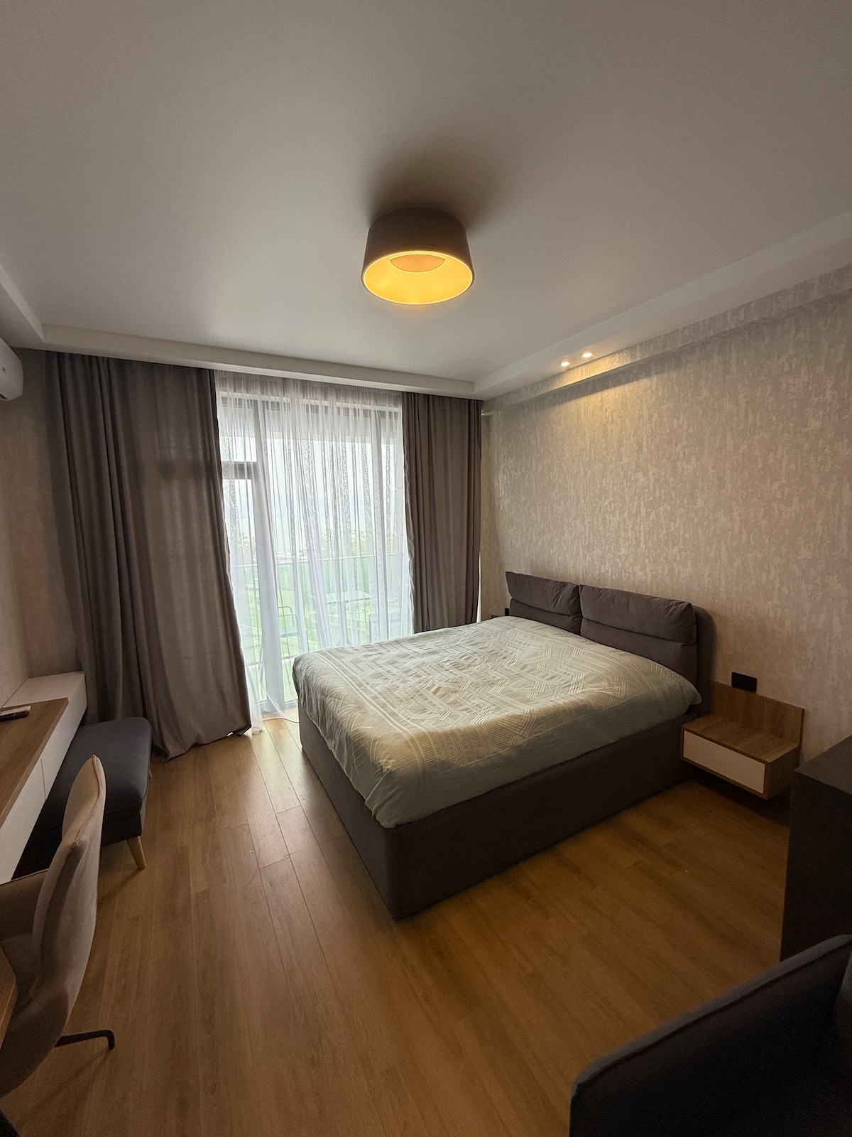 A bright bedroom features a spacious bed with a light-colored quilt. Floor-to-ceiling windows are dressed in sheer curtains, allowing natural light to enter. A small bedside table and a chair with a desk are positioned against the wall, complemented by warm wooden flooring.