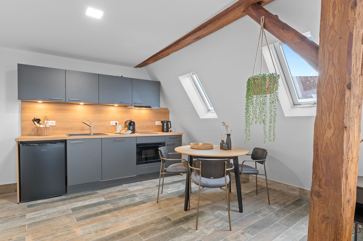 A modern kitchen area is equipped with dark cabinetry and wooden countertops, complementing the open space. Two skylights provide natural light, while a round dining table with four chairs is situated nearby, enhancing the functional layout. A potted plant hangs near the window.