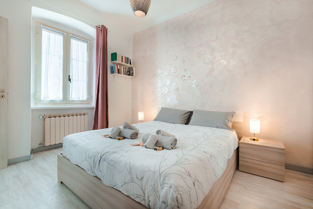 A spacious bedroom features a large bed with simple bedding and two nightstands, each with a lamp. Natural light filters through two windows adorned with sheer curtains. The decor includes subtle wall art and a small bookshelf, creating a serene atmosphere.