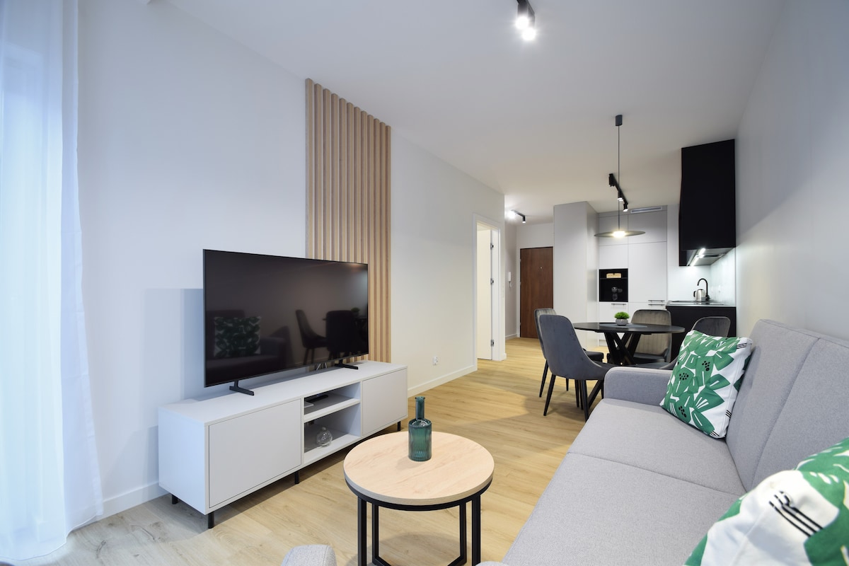 A contemporary living area features a light grey sofa and a wooden coffee table. A flat-screen television is positioned on a low white cabinet. Dining chairs surround a dark table in the background, and a well-equipped kitchen is visible, providing a cohesive and functional space.