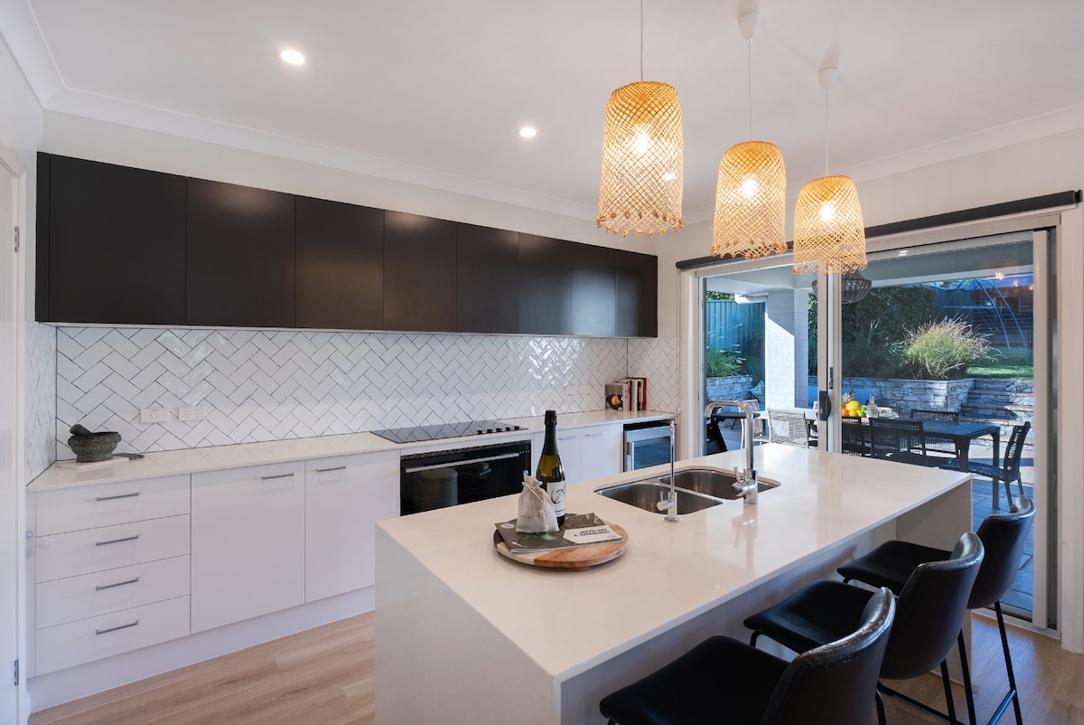 The modern kitchen features sleek cabinetry in dark tones paired with a light countertop. A spacious island includes a sink and seating area, while three woven pendant lights illuminate the space. Sliding glass doors open to an outdoor dining area, enhancing the inviting atmosphere.