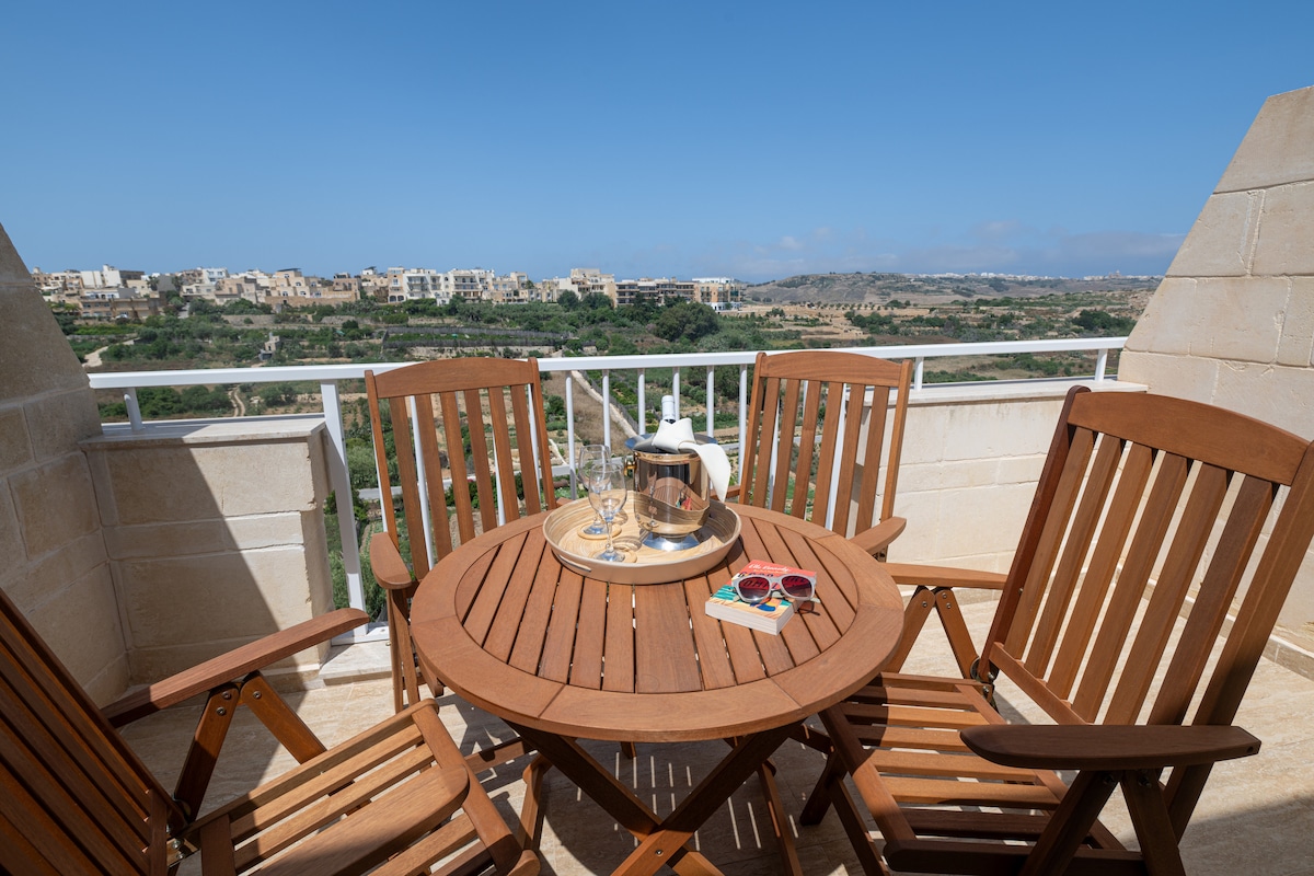 A wooden table is set on a balcony, surrounded by four matching chairs. A bottle of champagne is placed in an ice bucket, accompanied by two glasses and a pair of sunglasses. The expansive view of the valley and distant town is seen in the background.