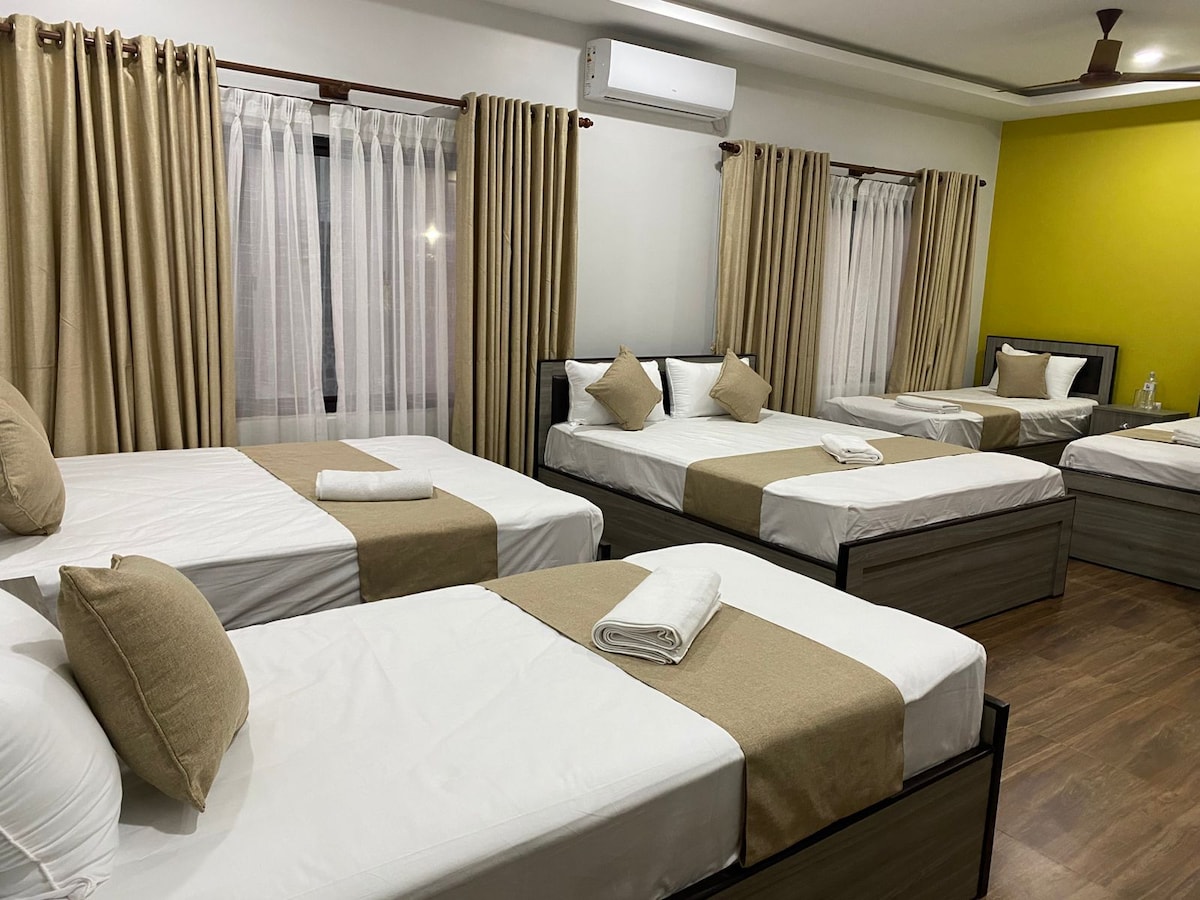 A spacious room is furnished with multiple beds, each featuring neutral-toned linens and soft pillows. Sheer white curtains allow natural light to filter in, while a vibrant green accent wall adds a pop of color. Air conditioning is visible on the ceiling.