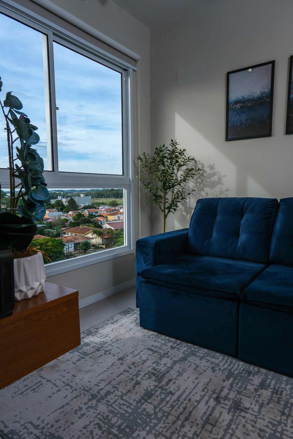 A cozy living area is presented with a plush blue sofa positioned near a large window. The view showcases a cityscape with greenery. Decorative plants are placed beside the sofa, and framed artwork adorns the wall, enhancing the room's inviting ambiance.