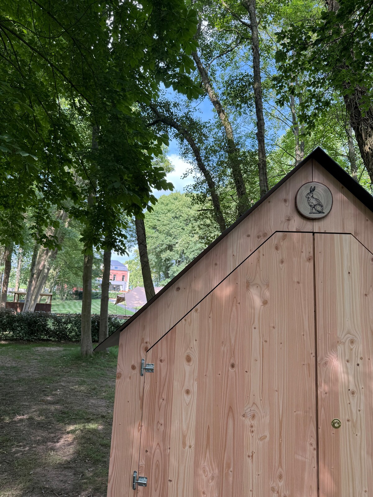 A wooden cabin is nestled among tall trees, with its natural wood exterior complementing the surrounding greenery. The sloped roof is detailed with a circular emblem. Soft sunlight filters through the leaves, creating a serene atmosphere in the natural setting.