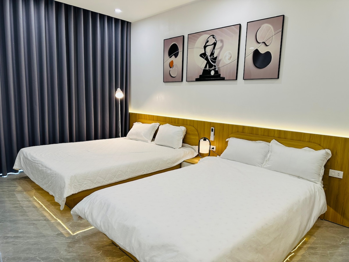 The sleeping area includes two spacious beds with crisp white linens, set against a modern backdrop of neutral tones. Artwork adorns the wall above, while dark curtains frame a window, providing a cozy and serene ambiance.
