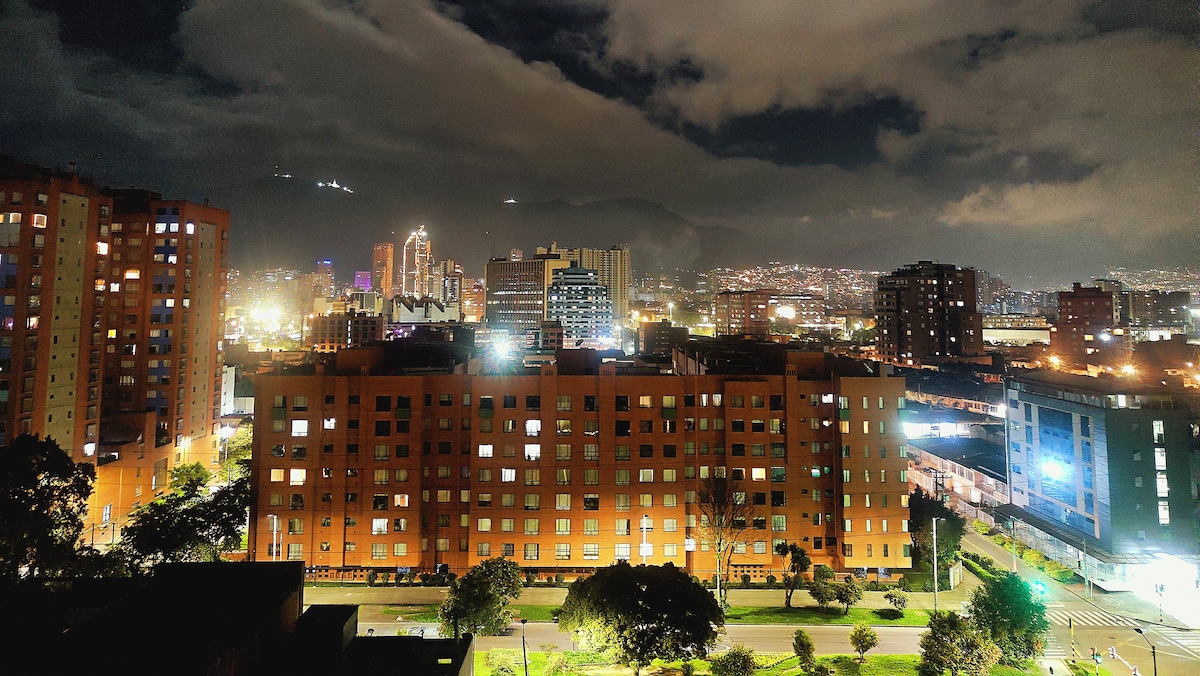 A cityscape is captured at night, featuring illuminated buildings and a vibrant skyline. The foreground displays a row of orange apartment blocks, while the background reveals distant lights of the city, under a cloudy sky. The ambiance reflects a lively urban atmosphere.