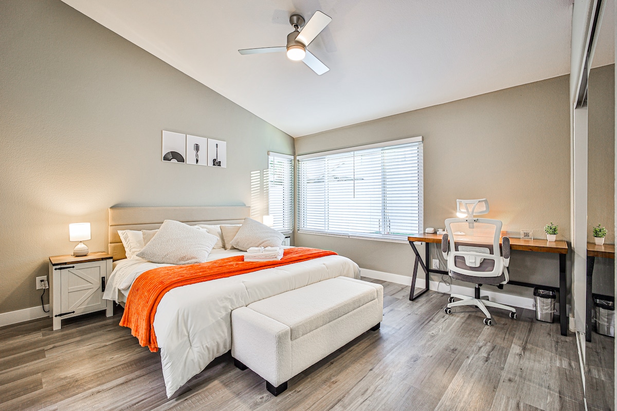 Werk comfortabel aan het bureau in de grote slaapkamer van The Vegas Hideout, met een licht houten bureau, een ergonomische witte stoel en natuurlijk licht. Een kingsize bed met wit beddengoed en een oranje deken voegt stijl toe aan deze rustige, uitnodigende ruimte.