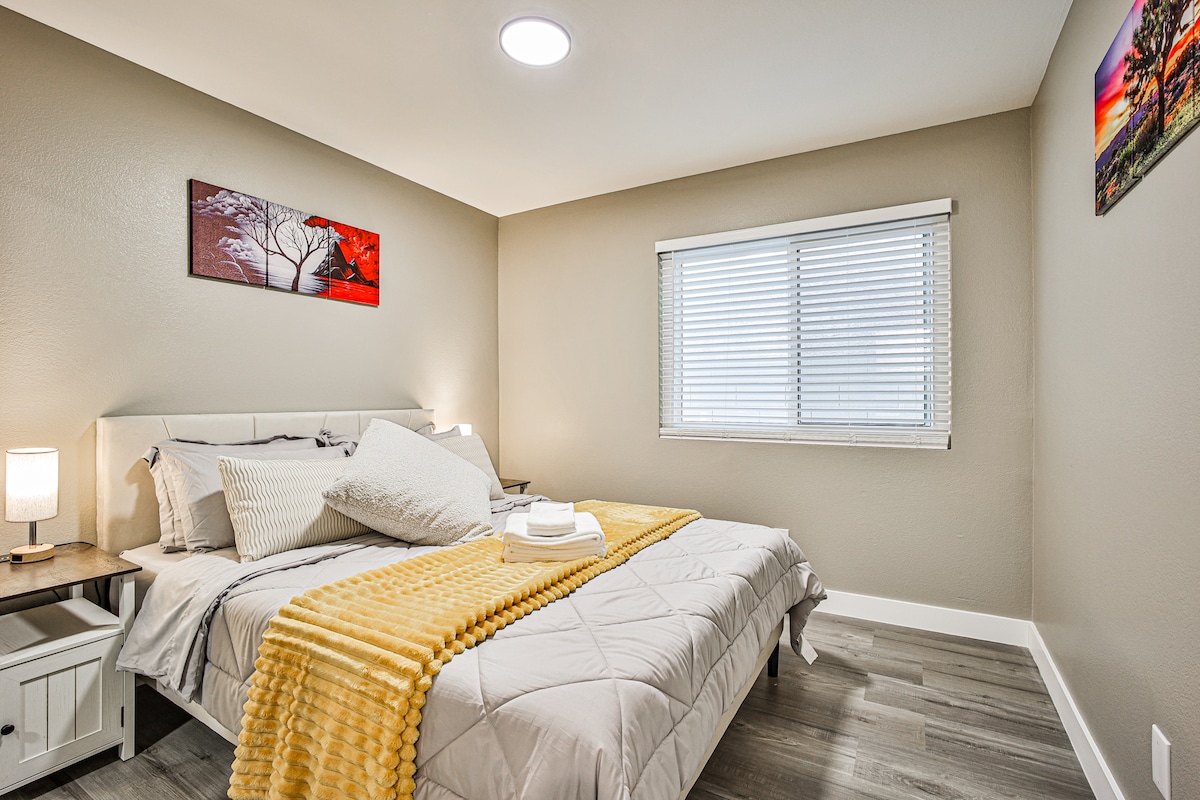 Ontspan in deze serene Vegas Hideout-slaapkamer met een queensize bed met grijs beddengoed, mosterdkleurige plaid, gelaagde kussens en kunst met woestijnthema. Lichte houten vloeren, zachte verlichting en witte jaloezieën zorgen voor een rustige, uitnodigende sfeer.