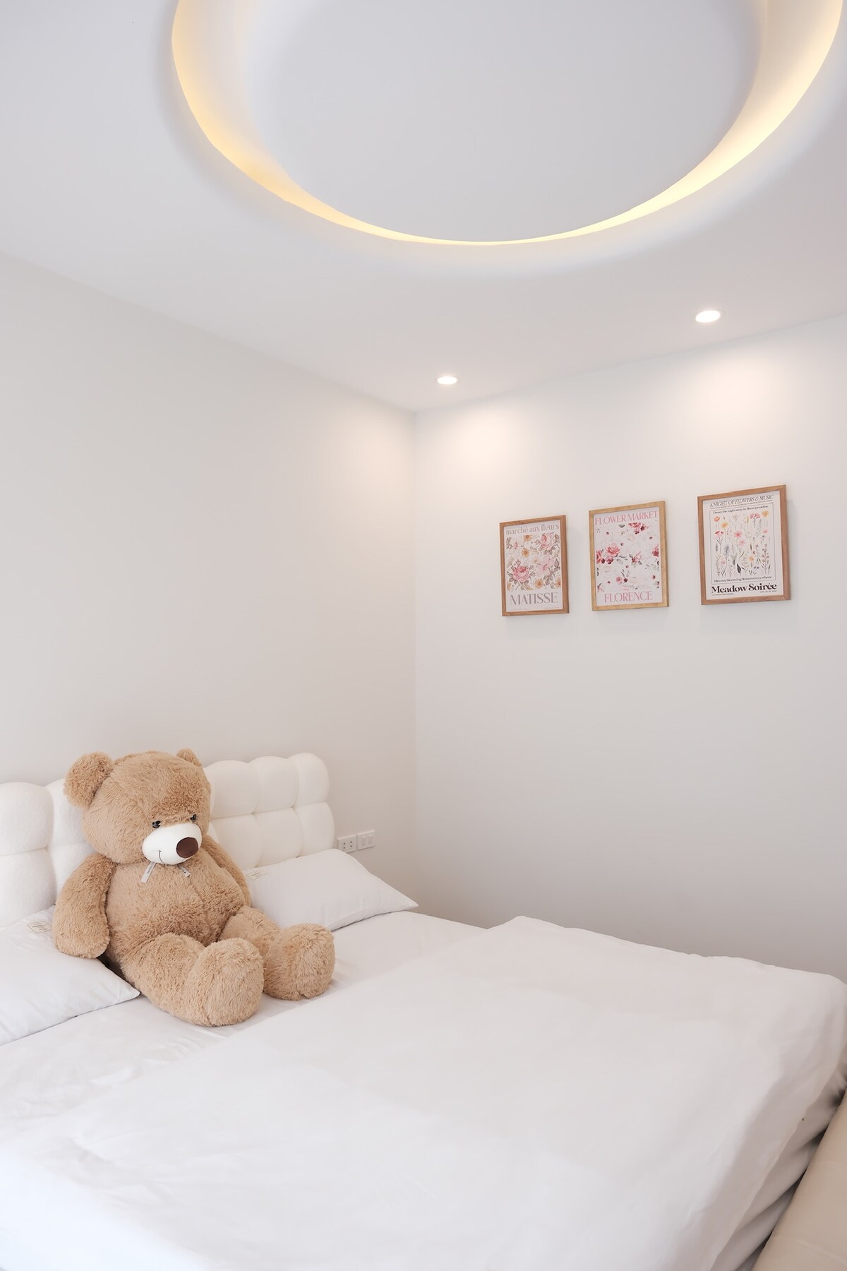 A spacious bed is adorned with soft white linens. A large teddy bear sits comfortably in the center, adding a touch of warmth. Three framed artworks are displayed on the light gray wall, providing a subtle decorative element. Soft lighting enhances the calming ambiance.