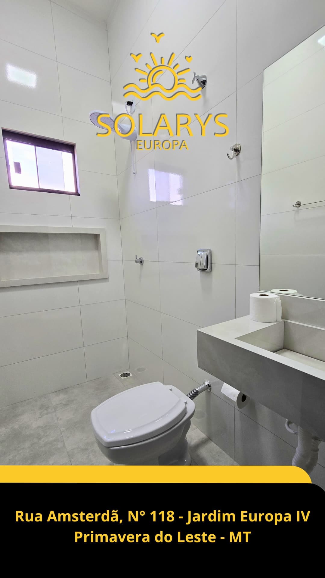 The bathroom features modern fixtures with a clean design. A wall-mounted sink is accompanied by a rectangular mirror. A flush toilet is positioned beside a small window that allows natural light to enter, enhancing the space's overall brightness.