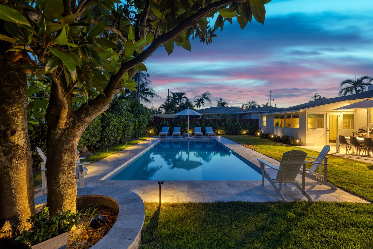 A serene backyard features a saltwater pool surrounded by lush green grass and elegant landscaping. Two mature trees provide shade, and several sun loungers are positioned by the pool. Soft lighting enhances the tranquil atmosphere as the sky displays a colorful sunset.