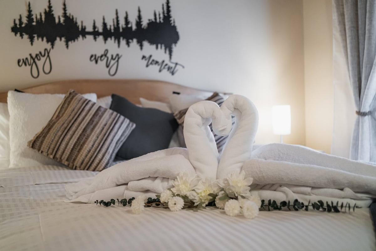 A neatly arranged bed is adorned with soft white linens and decorative pillows in shades of grey and beige. Towel swans form a heart shape at the center, accompanied by a decorative arrangement of flowers below. A bedside lamp casts a warm glow.