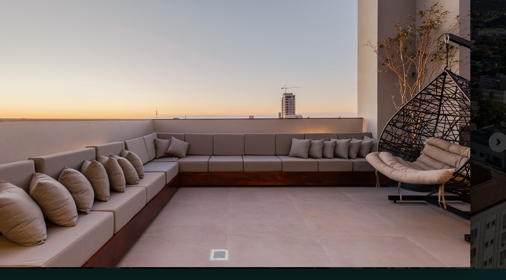 A spacious outdoor terrace presents a modern seating area with a large sectional sofa featuring soft cushions. A hanging chair is positioned in one corner, and the backdrop captures a serene sunset over the city skyline.