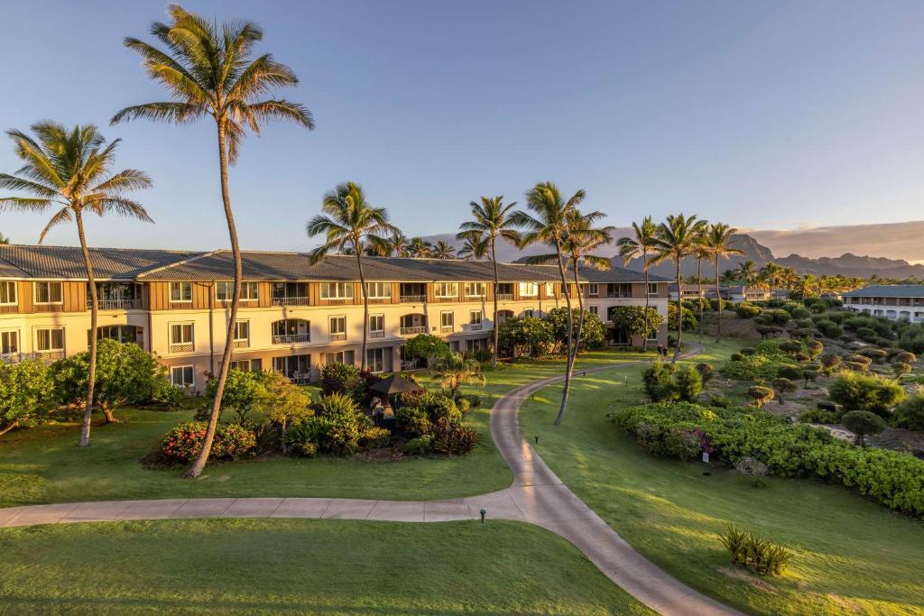 The image captures a resort area bordered by lush greenery and palm trees. A well-maintained pathway winds through the grounds, leading to multilevel accommodations. Spacious lawns are arranged throughout the landscape, creating a serene atmosphere with beautiful mountain views in the background.