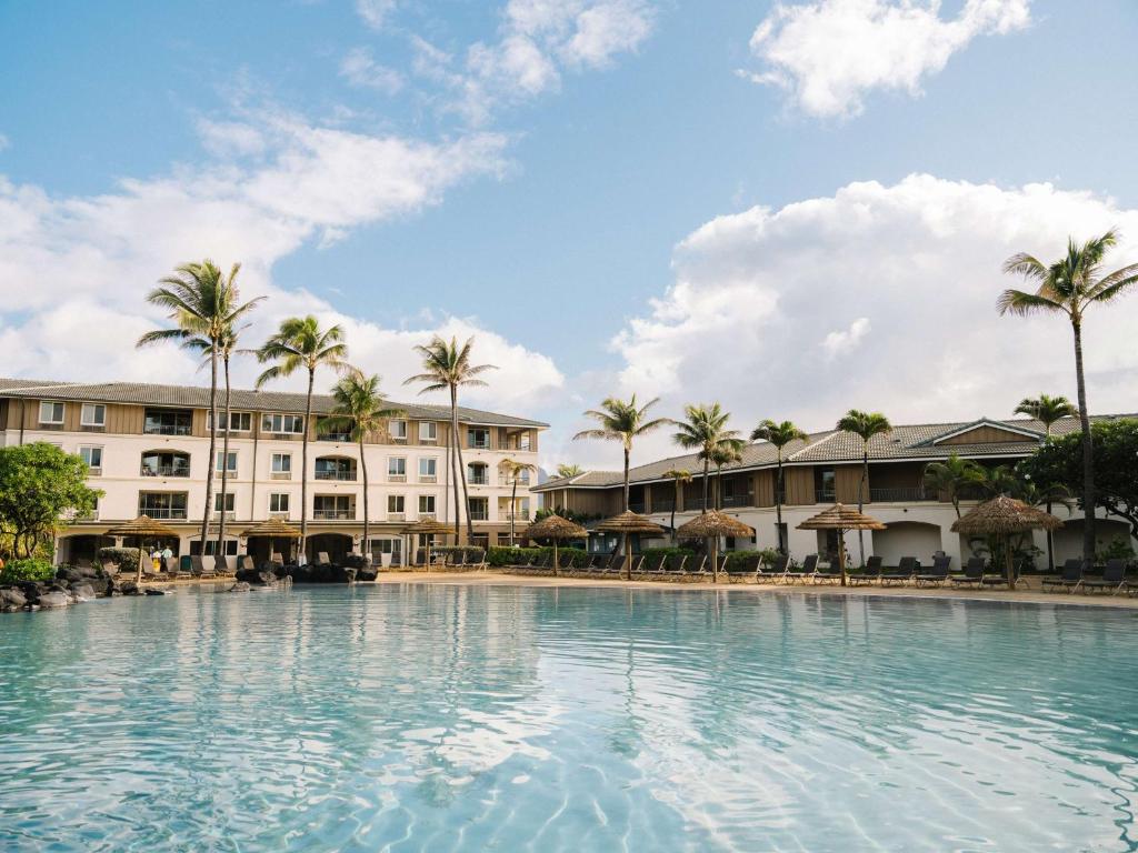 A serene resort pool is framed by palm trees and tropical landscaping. The building features multiple balconies and windows, suggesting spacious accommodations. Umbrellas shade lounge chairs by the pool, providing areas for relaxation under the Hawaiian sun.