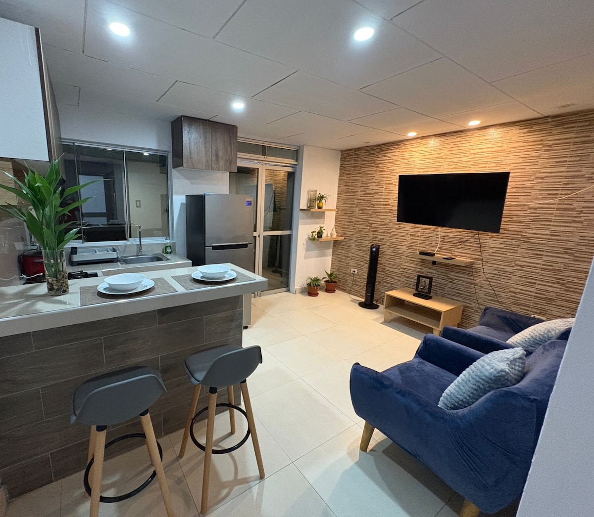 A contemporary kitchen is combined with a living area, featuring light-colored cabinets and a stainless-steel refrigerator. Comfortable seating is provided by plush blue armchairs, and a wall-mounted TV is visible above a sleek entertainment unit. Natural light filters through large sliding doors.