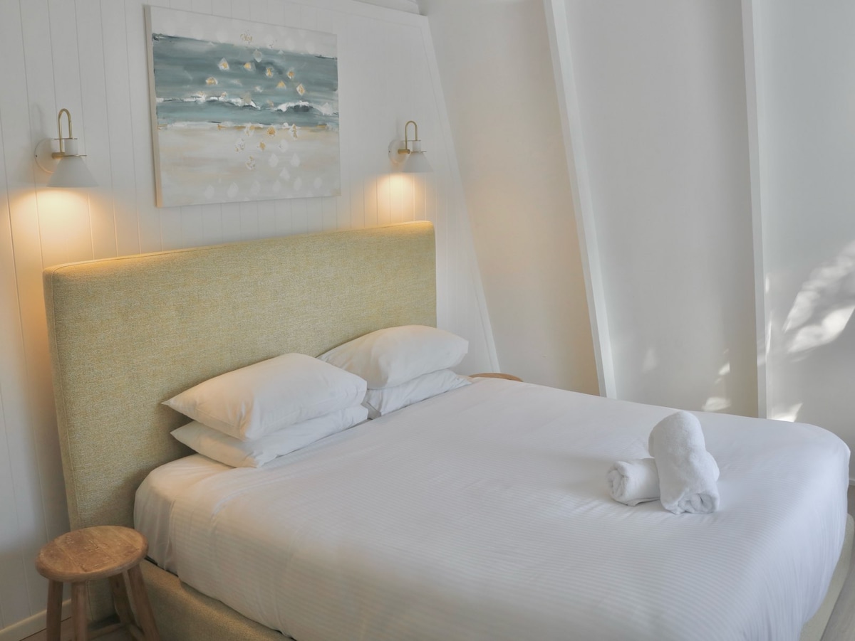 Spálňa 1 – slnečná miestnosť (hore, manželská posteľ)
Podkrovný apartmán orientovaný na severovýchod s bielymi drevenými trámami a čistou bielizňou. V tomto pokojnom pobrežnom útočisku sa nechajte unášať zvukmi oceánu.
