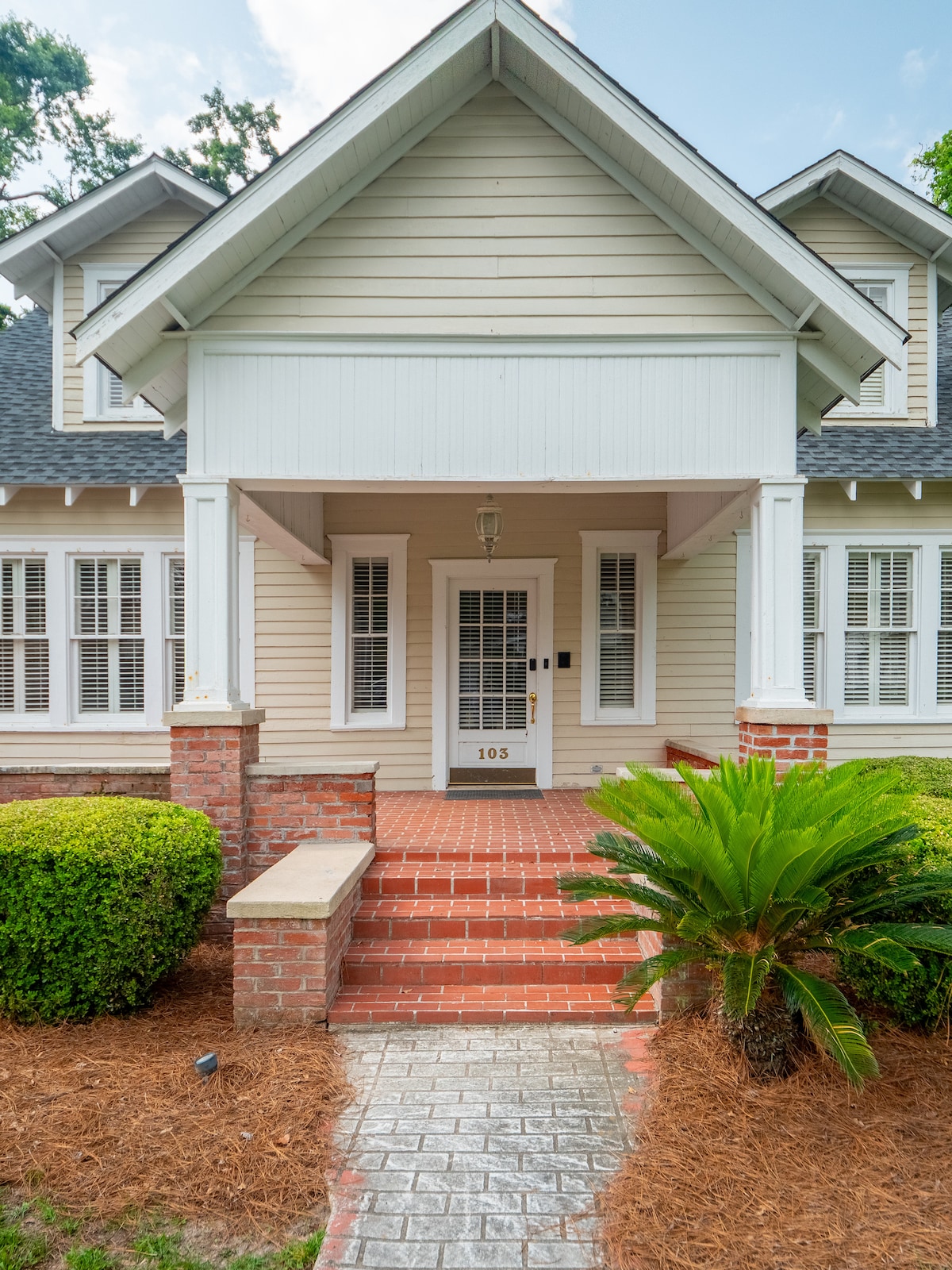 The front entrance of the historic home is framed by brick steps and vibrant greenery. A path of pavers leads up to the welcoming front door, flanked by large windows with shutters that enhance the home's architectural charm.