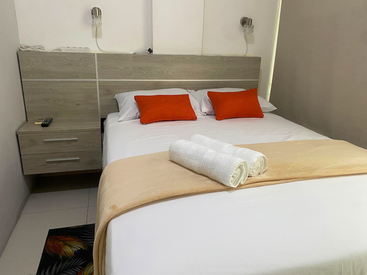 A spacious bed is in the center, adorned with soft white linens and two orange decorative pillows. A light beige throw is neatly placed at the foot of the bed. Simple nightstands are positioned on either side, complemented by modern wall lights.