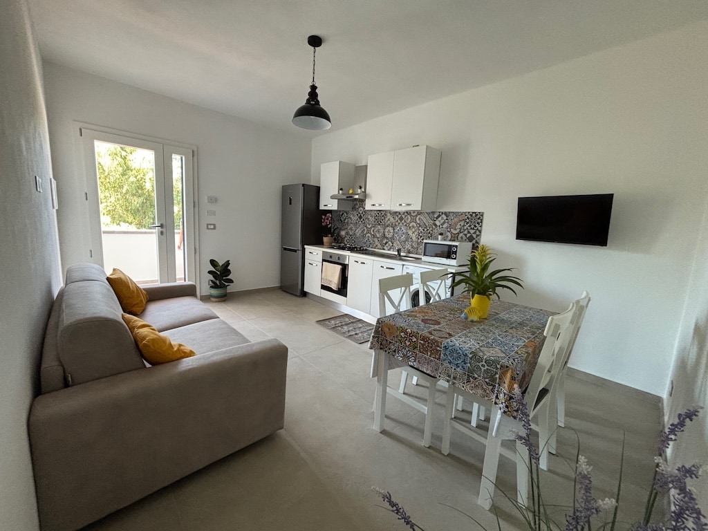 A modern living space features an open-plan kitchen and dining area, including a table with four chairs and a light-colored sofa. A wall-mounted television is visible. Natural light enters through the sliding glass door, which opens to a small outdoor area with greenery.