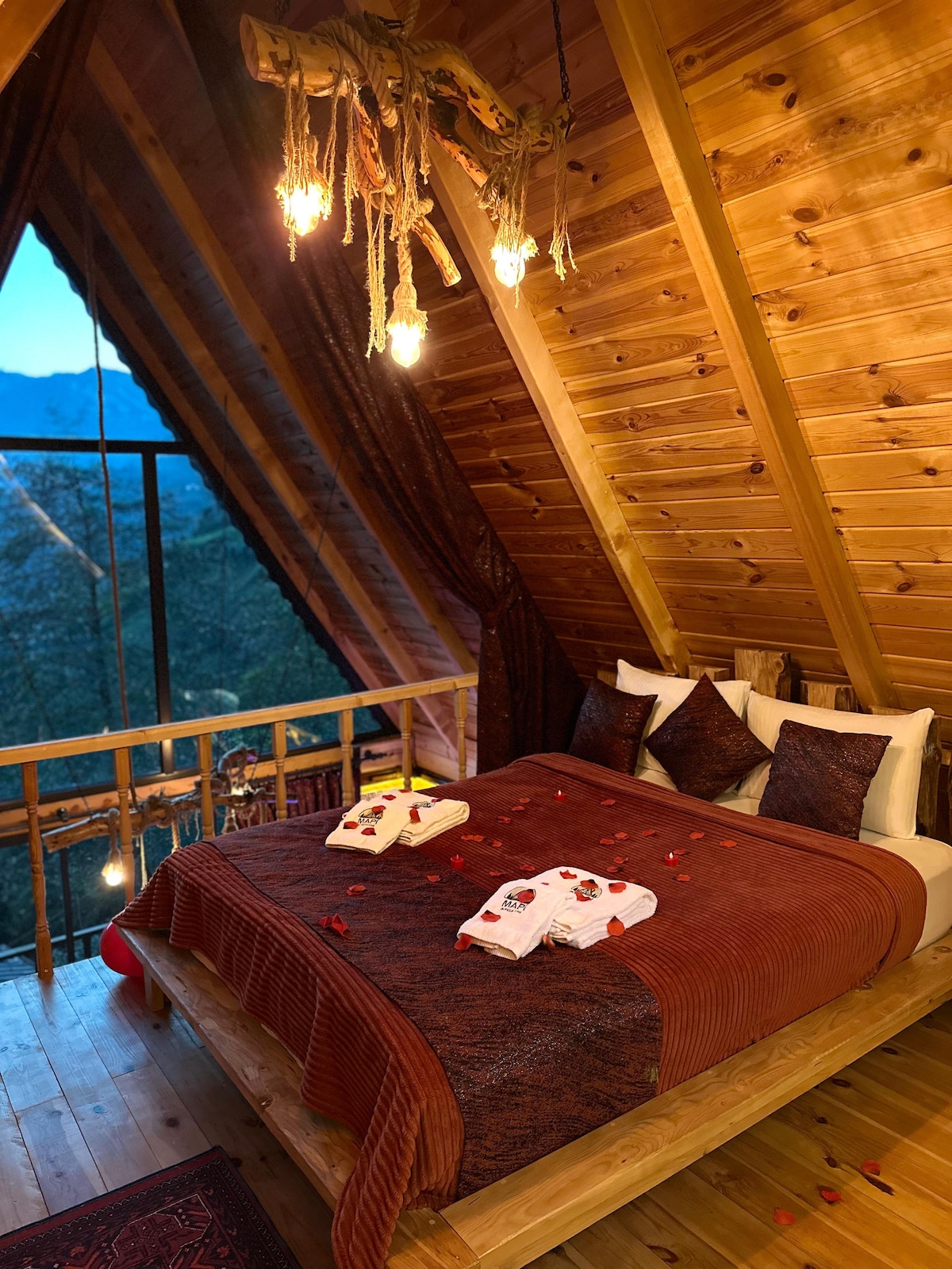 A spacious upper floor features a king-size bed adorned with decorative pillows and towels. Soft lighting from a rustic chandelier enhances the wooden interior, while large windows frame the surrounding mountain views. Rose petals are thoughtfully scattered across the bed, adding a romantic touch.