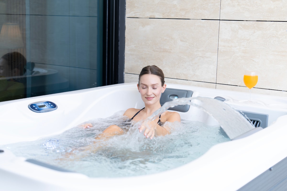 A private outdoor jacuzzi is showcased on the balcony, with water gently flowing from the side. A refreshing drink sits on the edge, complementing the serene atmosphere. The backdrop features modern wall tiles, creating a soothing environment for relaxation and enjoyment.