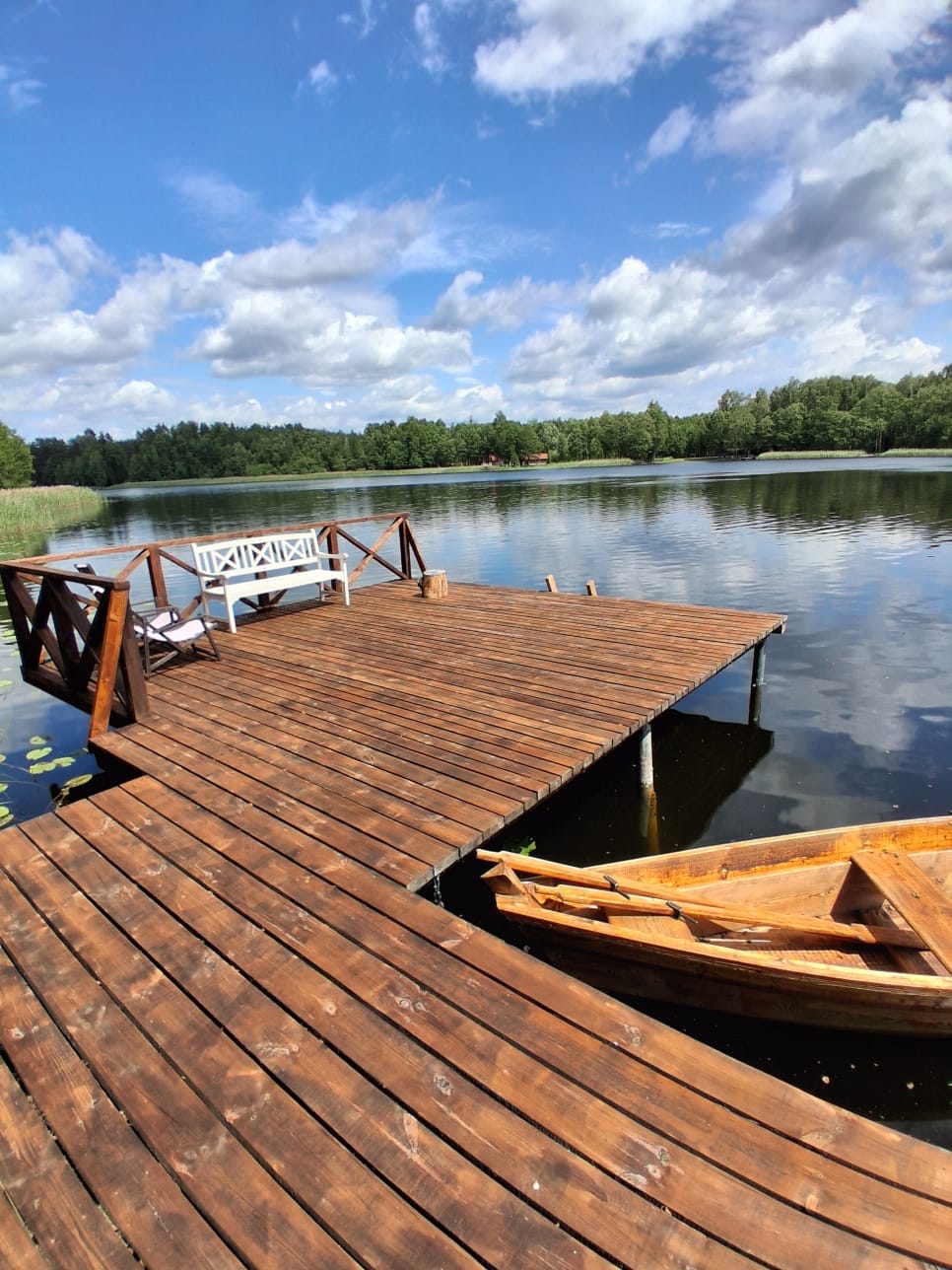 A wooden dock extends over calm waters, accompanied by a small boat tied at its edge. A white bench is positioned on the dock, offering a serene view of the surrounding nature and forested shoreline reflecting in the water.