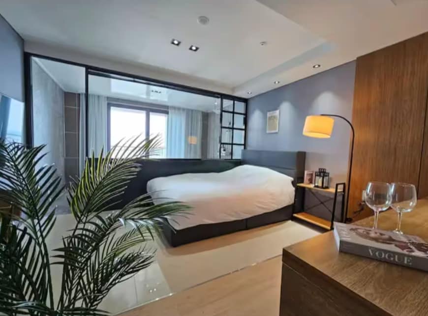 The suite features a spacious bed with a textured headboard, complemented by soft bedding. A large window allows natural light to fill the room. A small table holds wine glasses, and a floor lamp adds a modern touch. Decorative plants enhance the inviting ambiance.