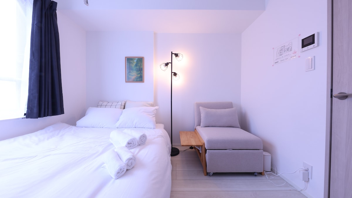 A well-lit sleeping area includes a spacious bed with soft linens and neatly arranged towels. A comfortable armchair sits beside a small wooden table, complemented by a standing lamp. A piece of artwork hangs on the wall, adding a touch of color to the serene space.