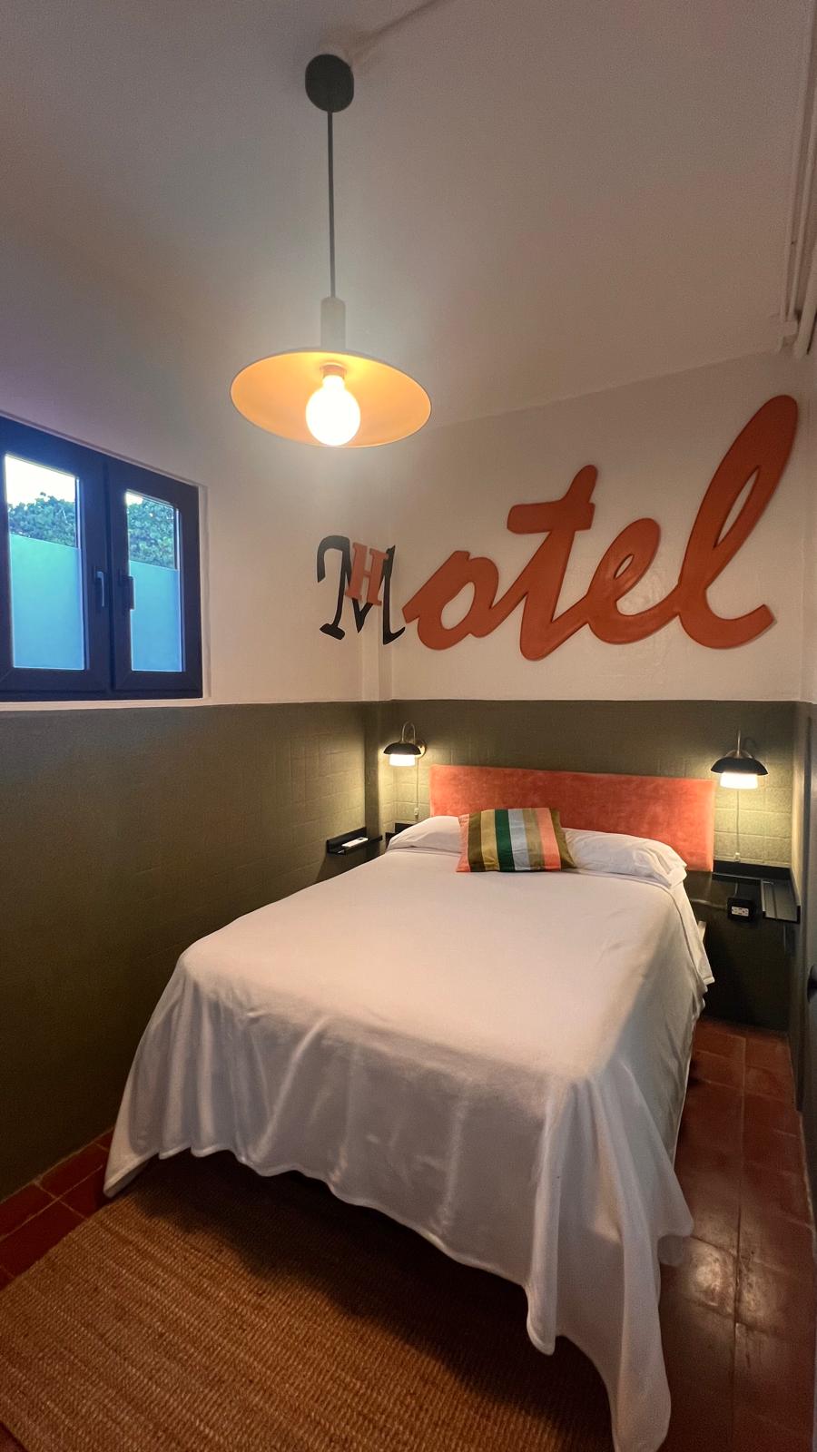 A cozy room is presented with a full bed covered by a white sheet. A large, stylized 'Motel' sign adorns the wall, complemented by two bedside lamps. A window allows for natural light, while a woven rug adds a touch of warmth to the tiled floor.