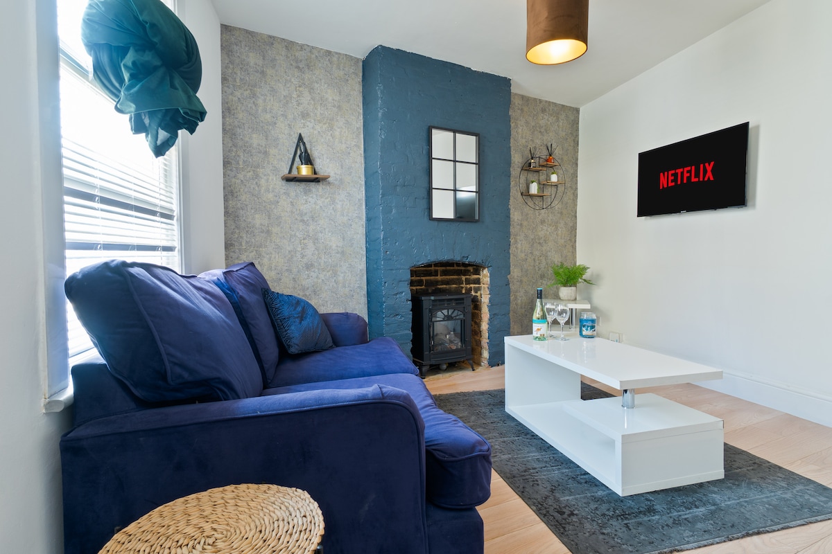 A cozy living area is highlighted by a navy blue sofa, complemented by a minimalistic coffee table and a textured area rug. The wall features a striking blue accent, with a television displaying the Netflix logo. Natural light streams in through the window, enhancing the welcoming ambiance.