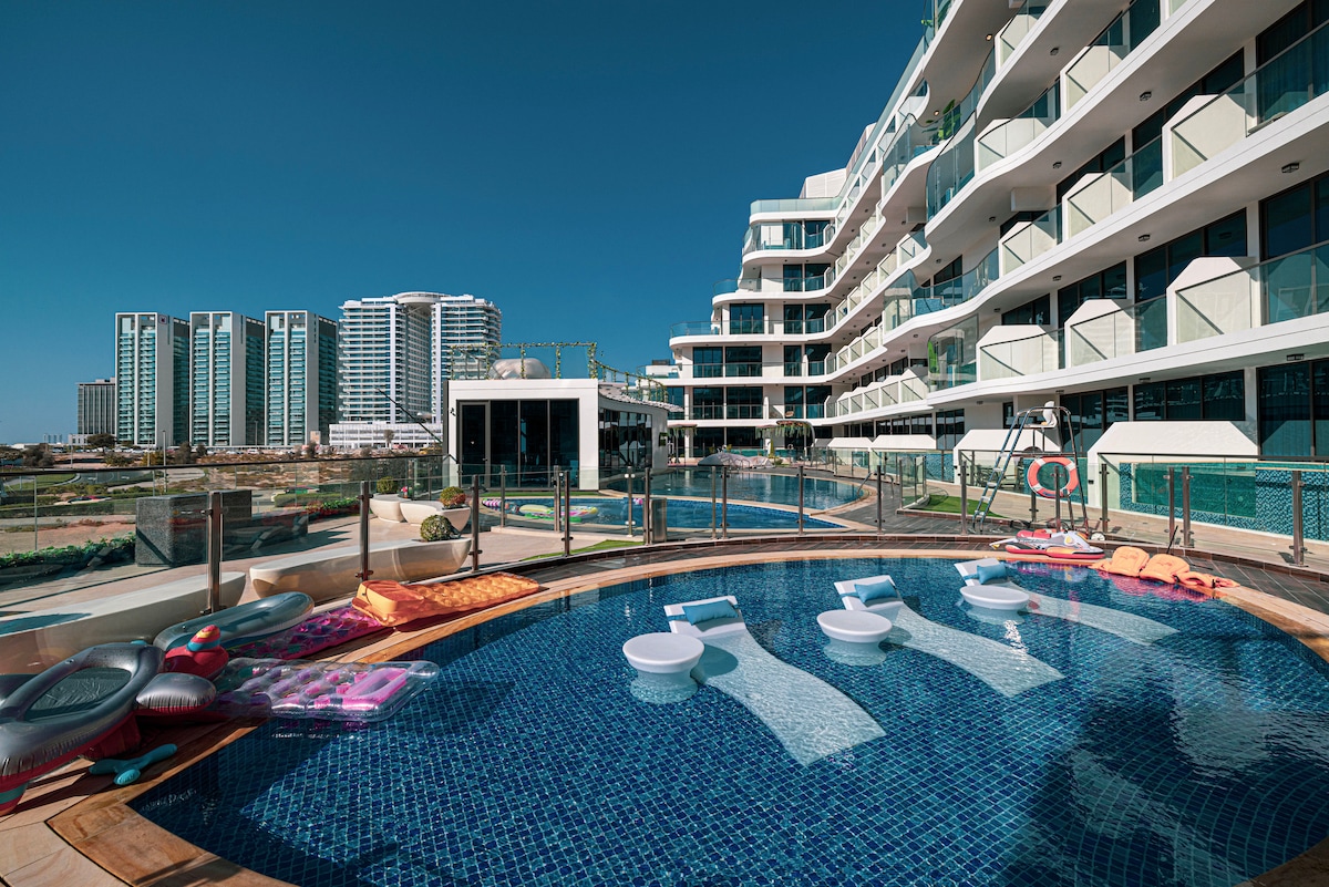 The serene terrace pool is surrounded by modern apartment buildings, featuring inviting seating options on the water's edge. Inflatable loungers and vibrant pool floats rest beside the shimmering water, creating a welcoming atmosphere for relaxation and leisure.