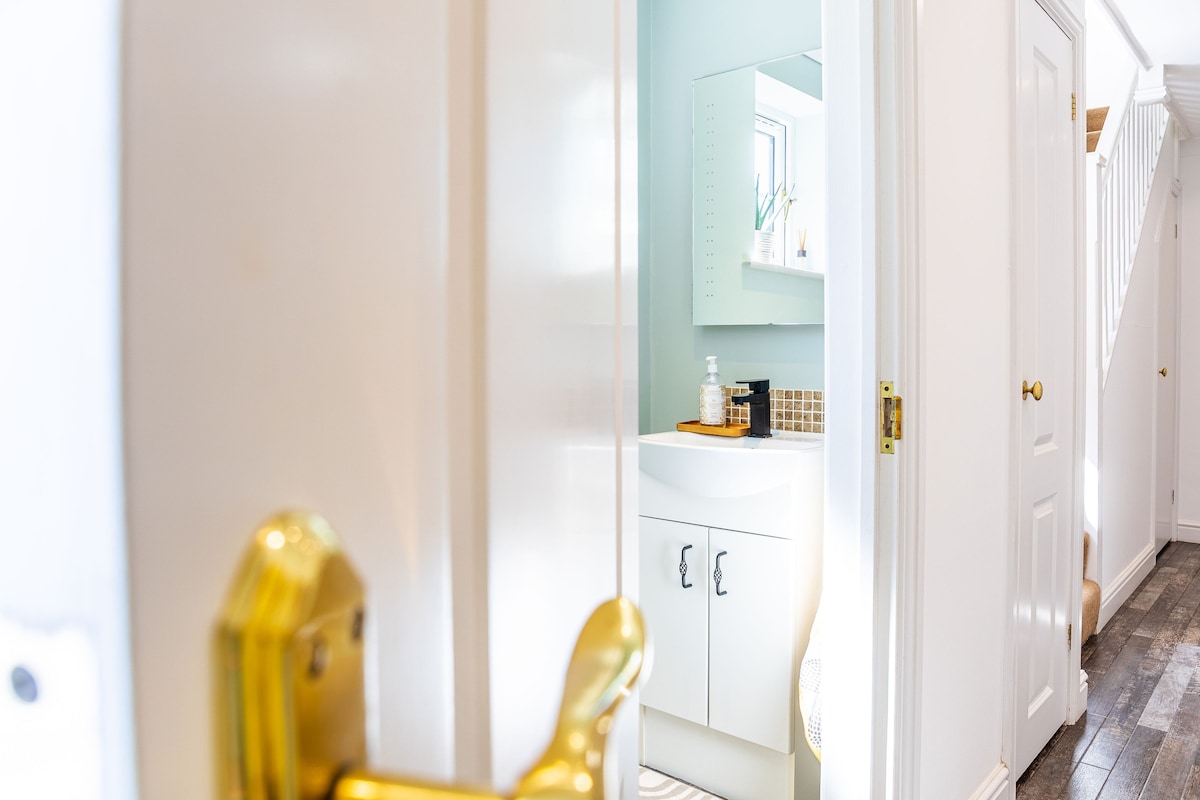 A well-lit bathroom is visible through an open door, featuring a sink with a white cabinet below and a mirror above. Light blue walls complement the space, while a small decorative item sits atop the countertop. Soft flooring extends into the hallway beyond.