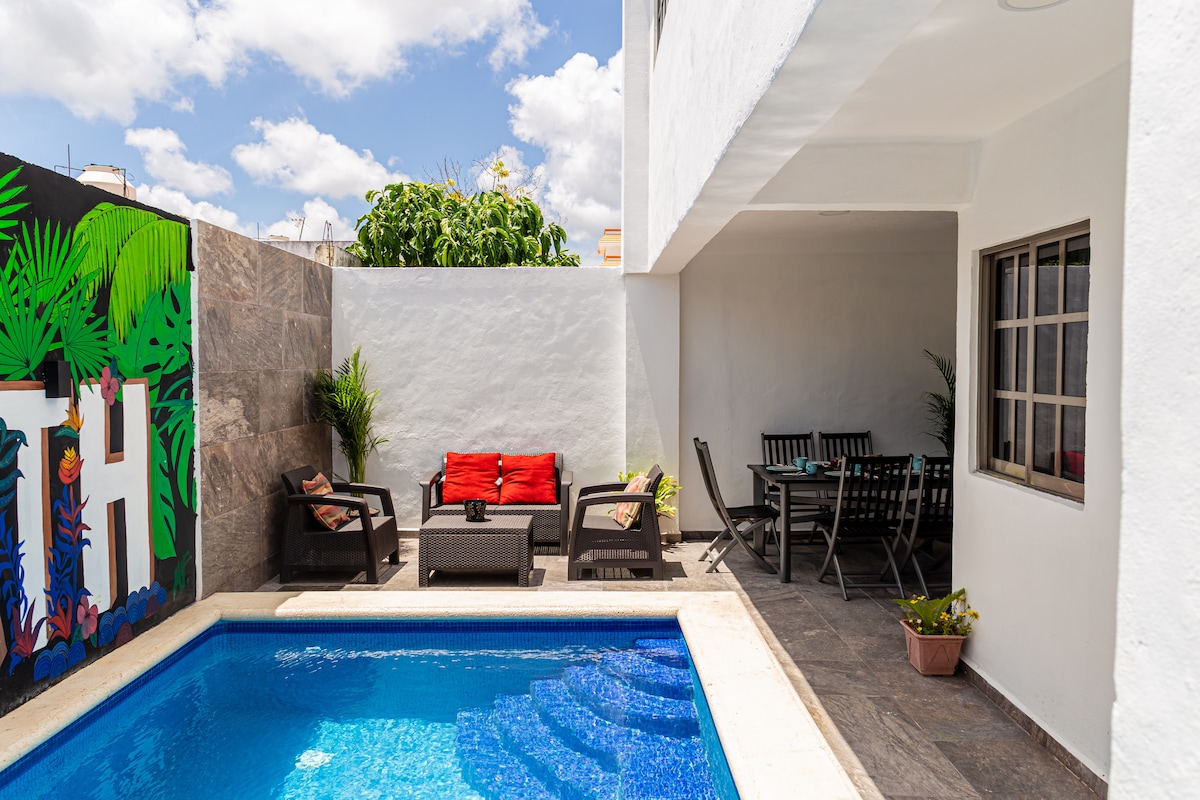 A private outdoor space features a swimming pool surrounded by a stone patio. Comfortable seating with cushions is arranged nearby, alongside a dining area with a table and chairs. Lush greenery enhances the serene environment, and colorful murals decorate the walls.