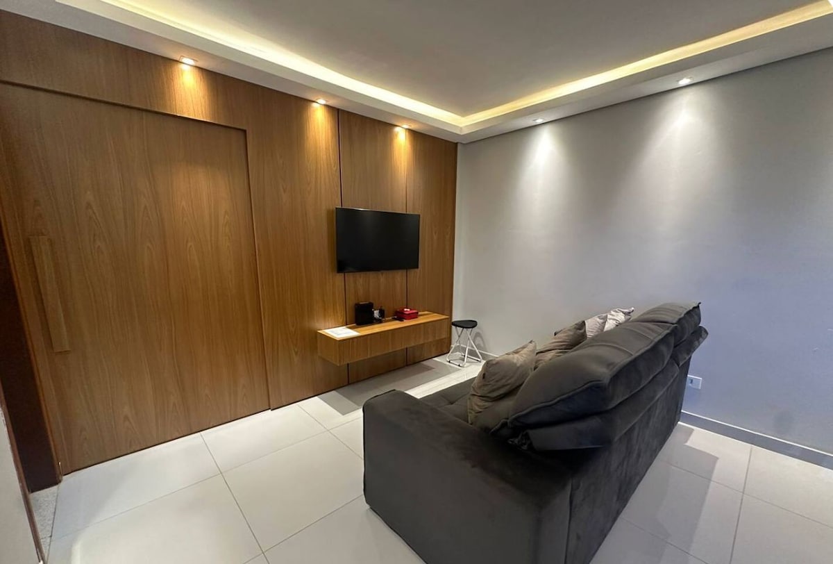 The living area features a comfortable, dark sofa facing a mounted television. A wooden panel wall adds warmth, and a sleek console table holds entertainment items. Soft lighting enhances the space's inviting atmosphere, while light tile flooring contributes to the modern aesthetic.