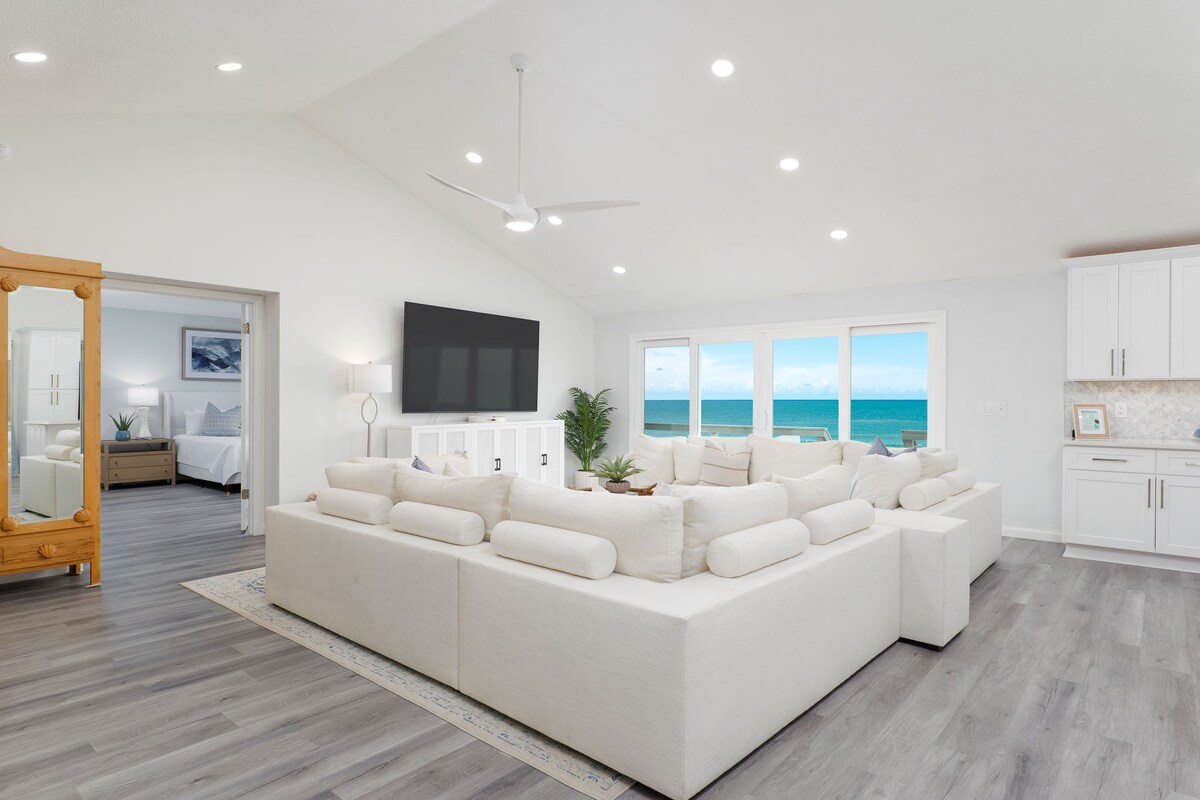 A spacious living room is highlighted by large windows providing ocean views. A sectional sofa offers ample seating, complemented by a large wall-mounted TV. Natural light illuminates the room, enhancing the modern design and airy atmosphere.