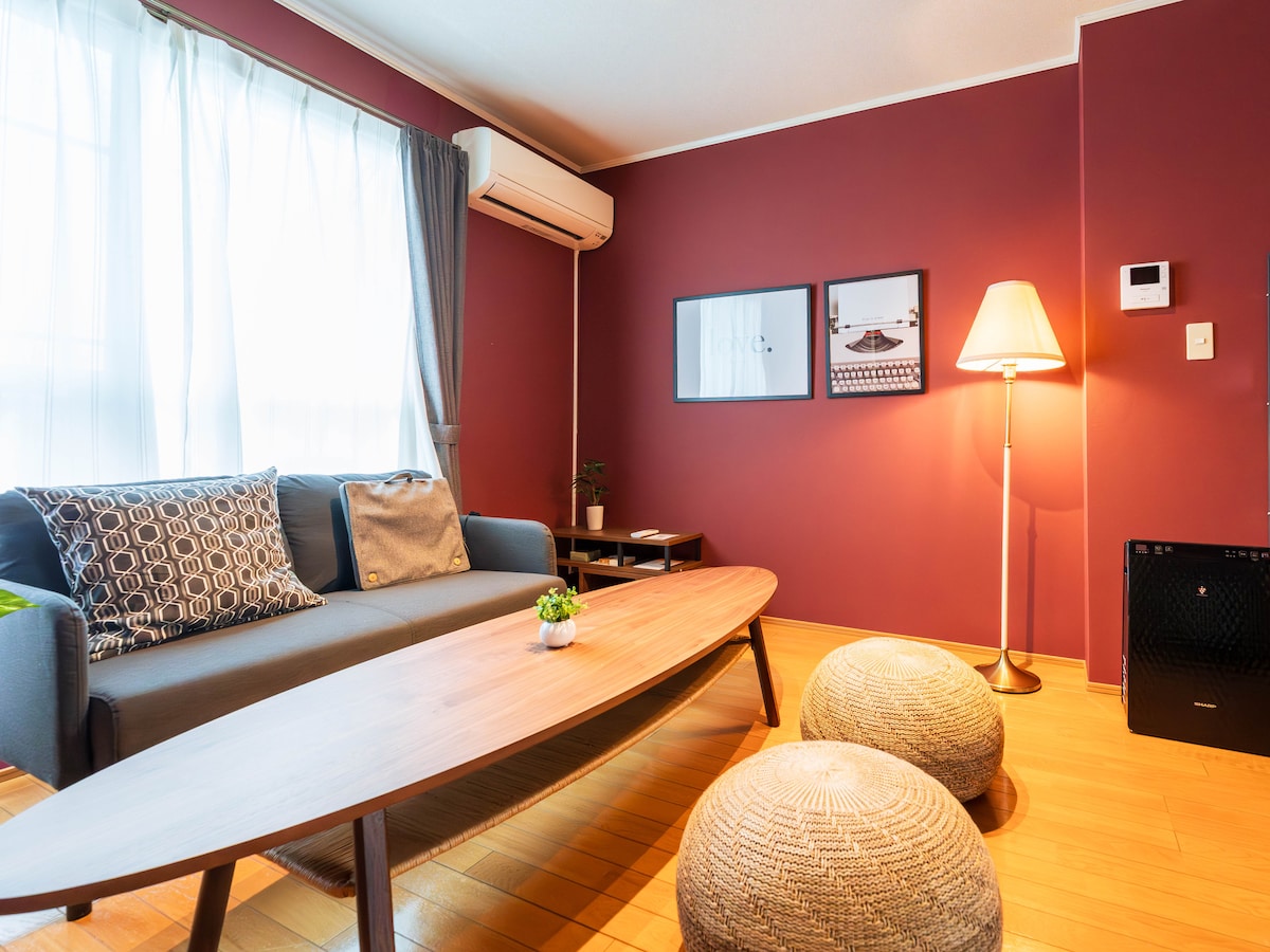 A living area features a comfortable sofa against a rich maroon wall. A stylish wooden table sits in the center, surrounded by two woven poufs. Natural light filters through sheer curtains, while framed photographs and a modern lamp add to the decor.