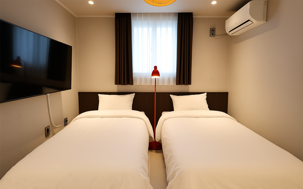 A well-lit guestroom features two spacious beds with crisp white linens. A modern television is mounted on the wall opposite the beds. Soft curtains frame the window, allowing natural light to fill the space, while a red lamp adds a pop of color.