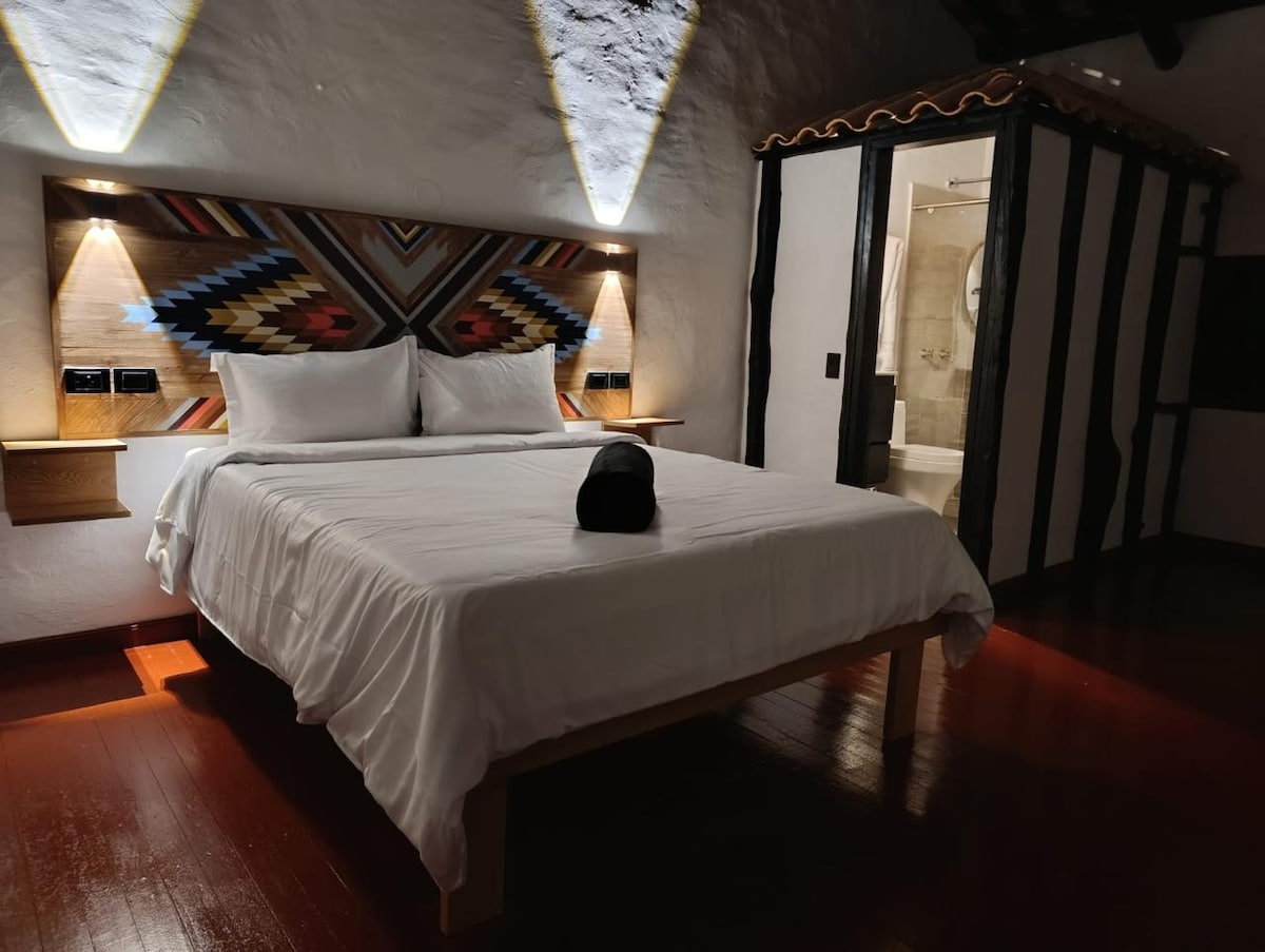 A spacious bed with white linens is positioned against a textured wall featuring geometric patterns. Soft lighting illuminates the bedside area. The room includes a wooden floor and visible access to a bathroom area through a decorative partition.