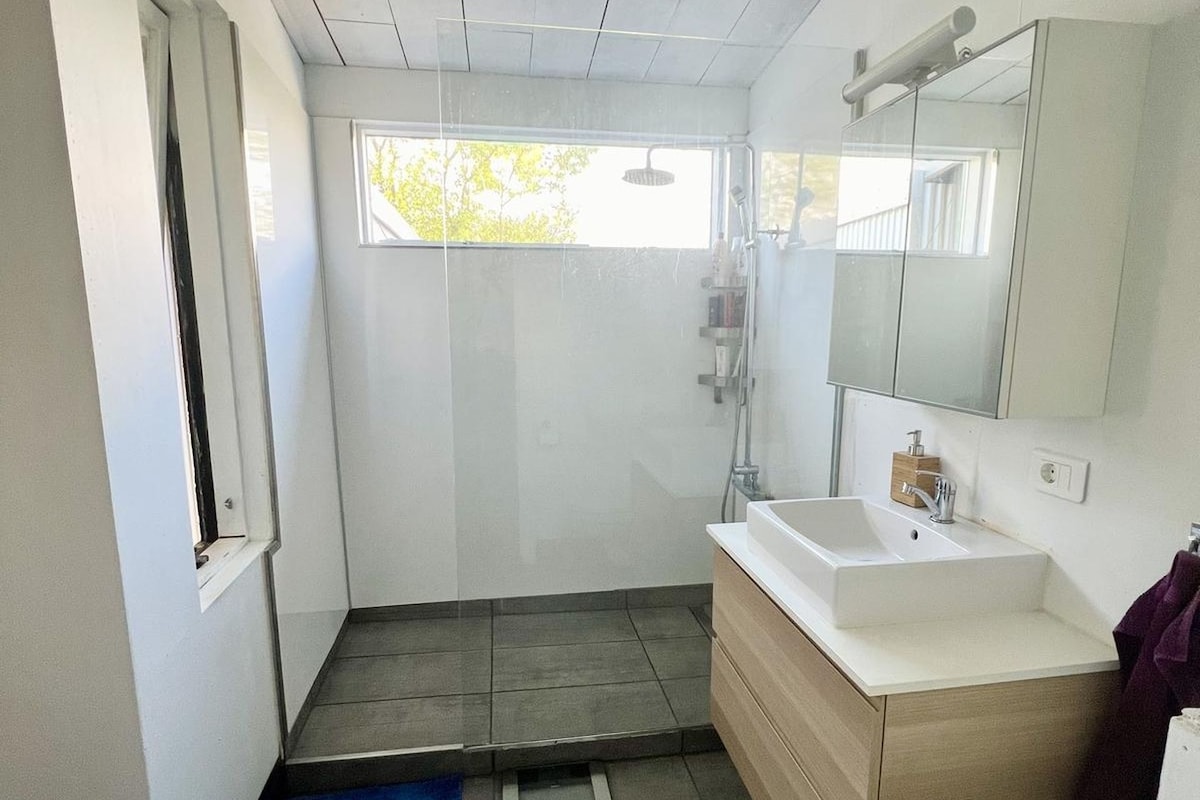 A modern bathroom features a spacious glass shower enclosed by sleek tiles. A vanity with a white sink is positioned against a light wooden cabinet. Natural light enters through large windows, creating a bright and open feel.