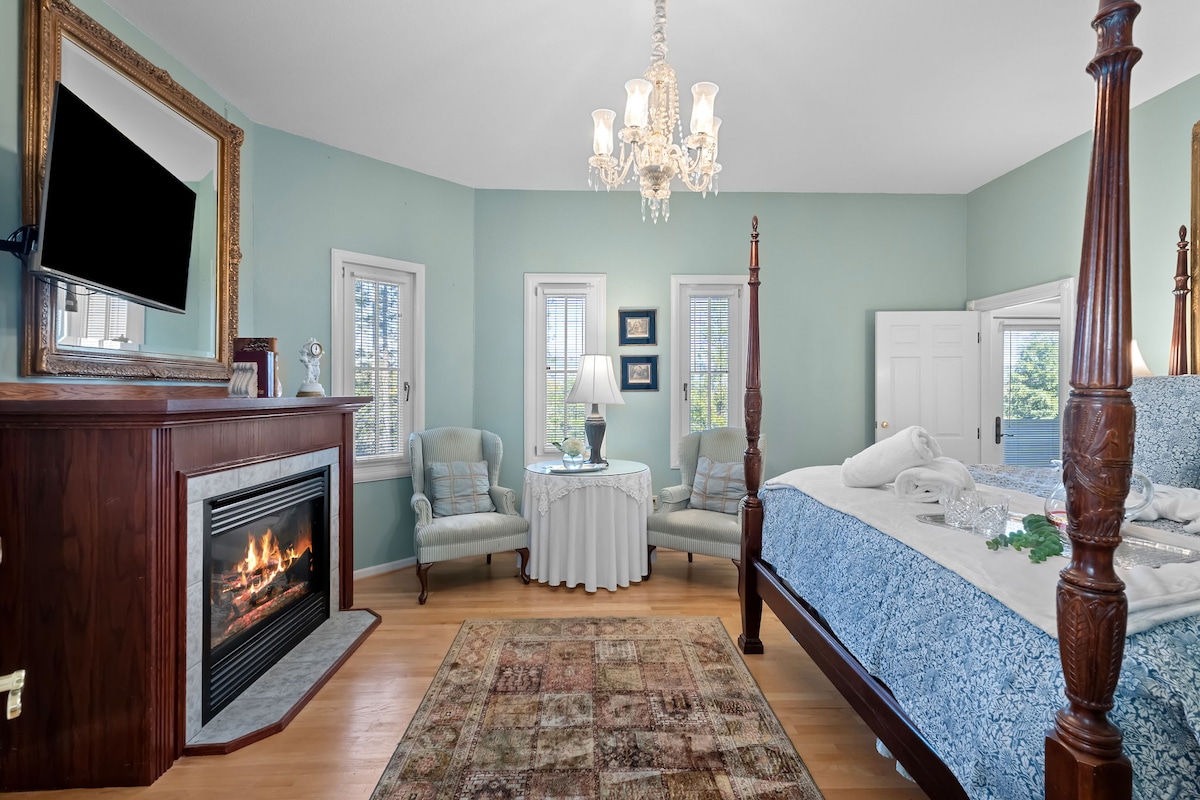 A well-appointed bedroom features a four-poster bed dressed in soft linens. A cozy seating area with elegant chairs is positioned next to a circular table. A decorative chandelier hangs above, while a fireplace adds warmth. Natural light filters through multiple windows.