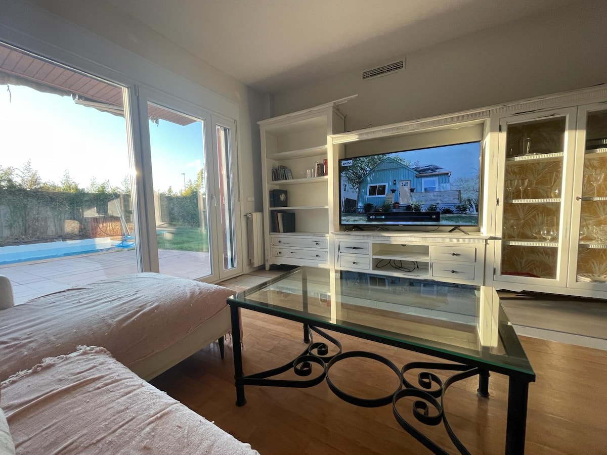 A bright living room is featured, with large sliding glass doors leading to the outdoor area. A spacious coffee table sits atop polished wood flooring, complemented by a comfortable sofa. A large television is mounted on a white shelf unit, providing ample storage space.