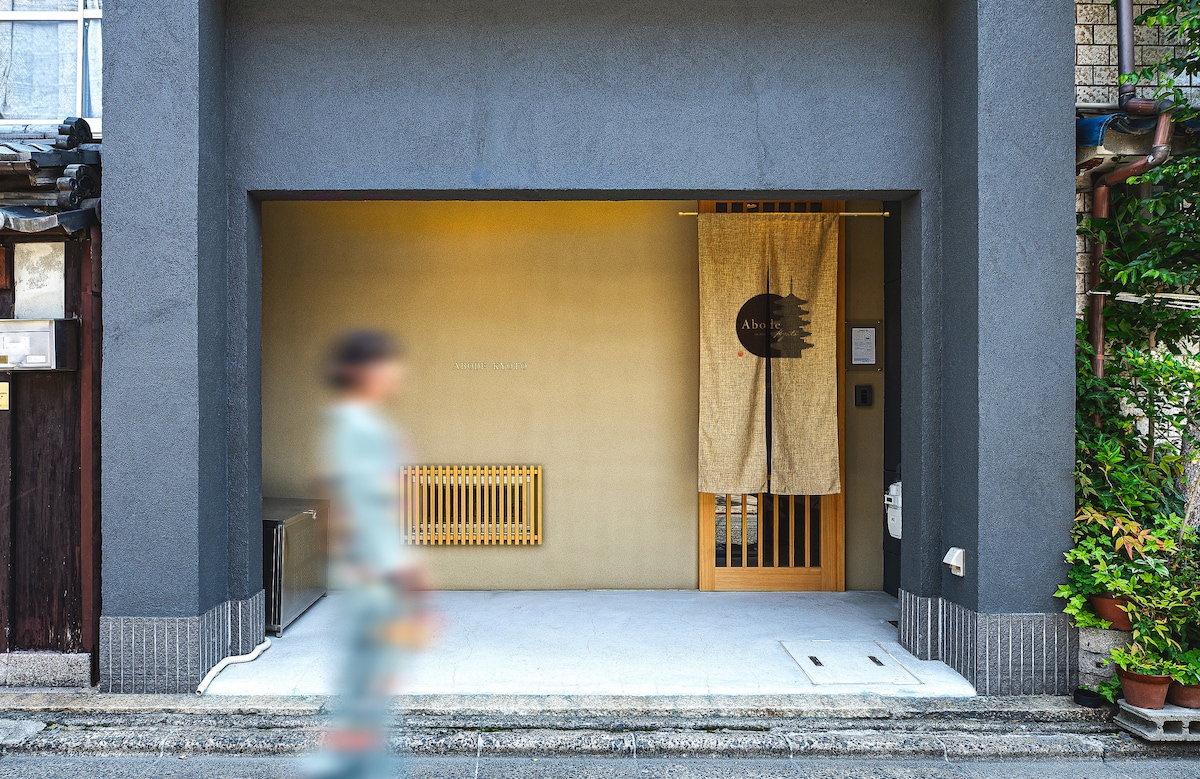 The entrance features a minimalist design with a light-colored wall and a subtle wooden accent. A decorative curtain hangs at the door, adding a touch of warmth. The clean, unobstructed entryway leads to the lodging, complemented by a smooth concrete pathway.